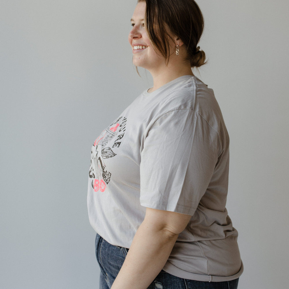 A woman in a "WHEN YOU'RE BAD AND BOOJEE" graphic tee by Love Marlow and blue jeans stands sideways, smiling, with her hair tied back.