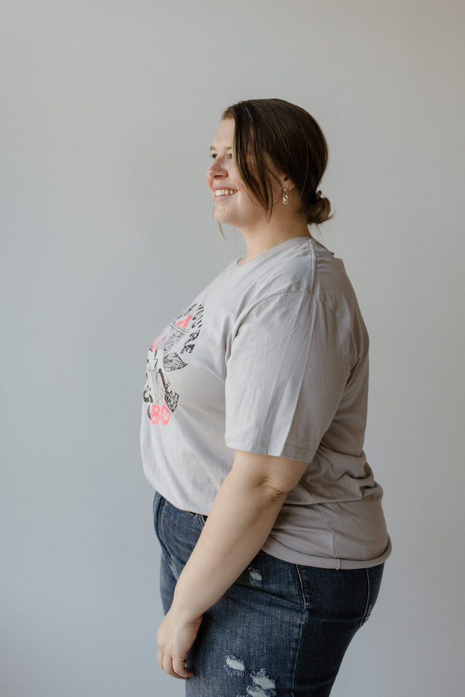 A person with brown hair in a bun wearing a "WHEN YOU'RE BAD AND BOOJEE" graphic tee by Love Marlow and jeans, facing away from the camera against a plain background, exudes understated bad and boojee vibes.