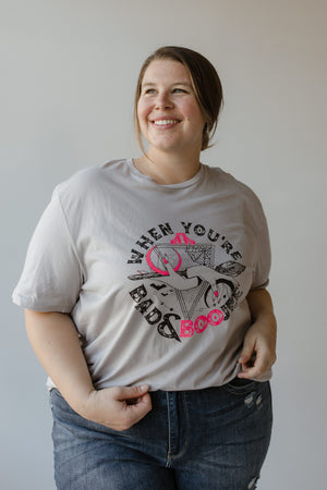 A person is smiling while wearing a graphic tee and jeans. The T-shirt, from Love Marlow, showcases a cowgirl hat, boots, and the phrase "WHEN YOU'RE BAD AND BOOJEE," making it perfect for capturing that bad and boojee vibe this Halloween season.