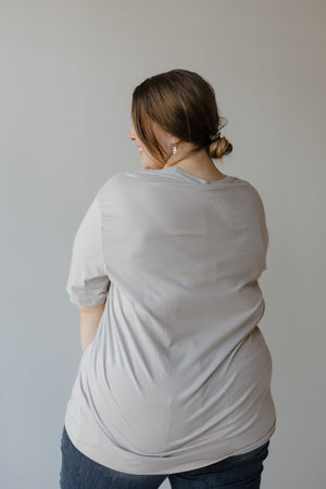 A person with brown hair in a bun wearing a "WHEN YOU'RE BAD AND BOOJEE" graphic tee by Love Marlow and jeans, facing away from the camera against a plain background, exudes understated bad and boojee vibes.