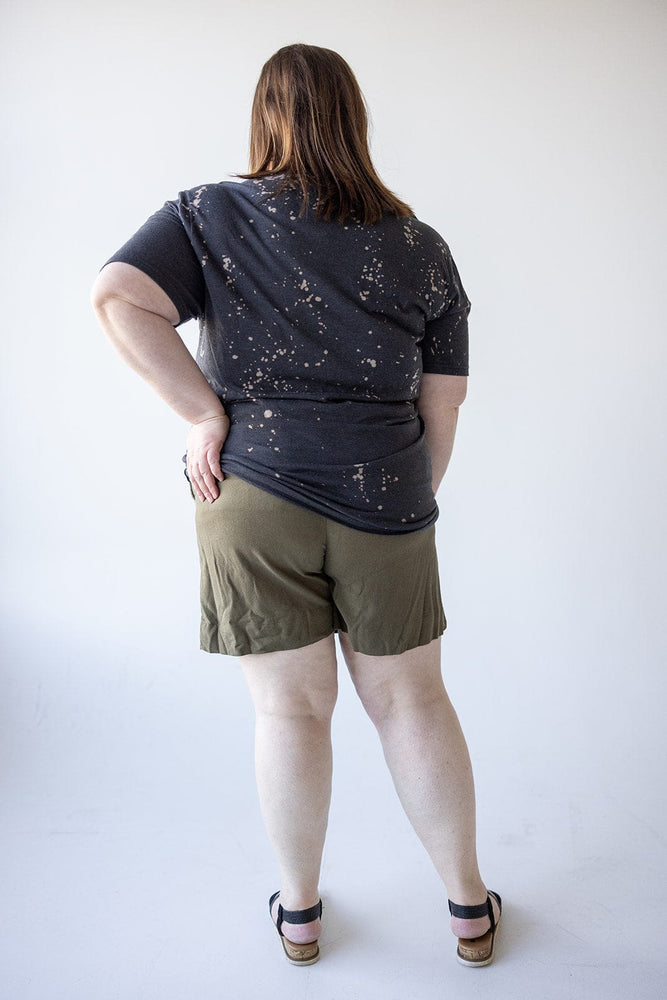 A person stands with their back to the camera, wearing a black, speckled shirt and WIDE LEG LINEN TROUSER SHORTS IN BERKEN by Dex, featuring an elastic waistband. Black sandals complete the look as they rest one hand on their hip.