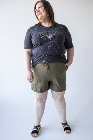 A person stands smiling in a casual outfit, wearing a dark gray graphic t-shirt and Dex WIDE LEG LINEN TROUSER SHORTS IN BERKEN with an elastic waistband, paired with black sandals.