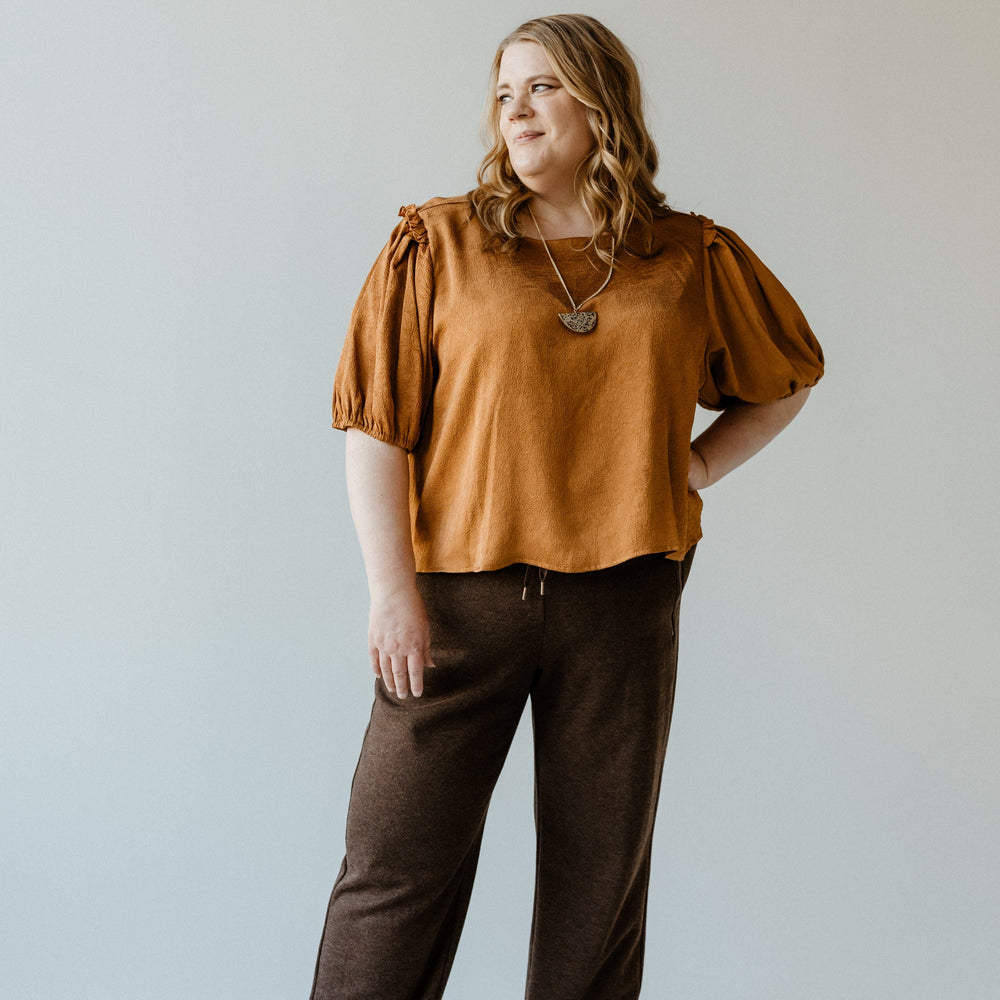 A person stands against a plain background, wearing a brown top and loose high-waist, wide-leg trousers paired with black shoes, looking off to the side.