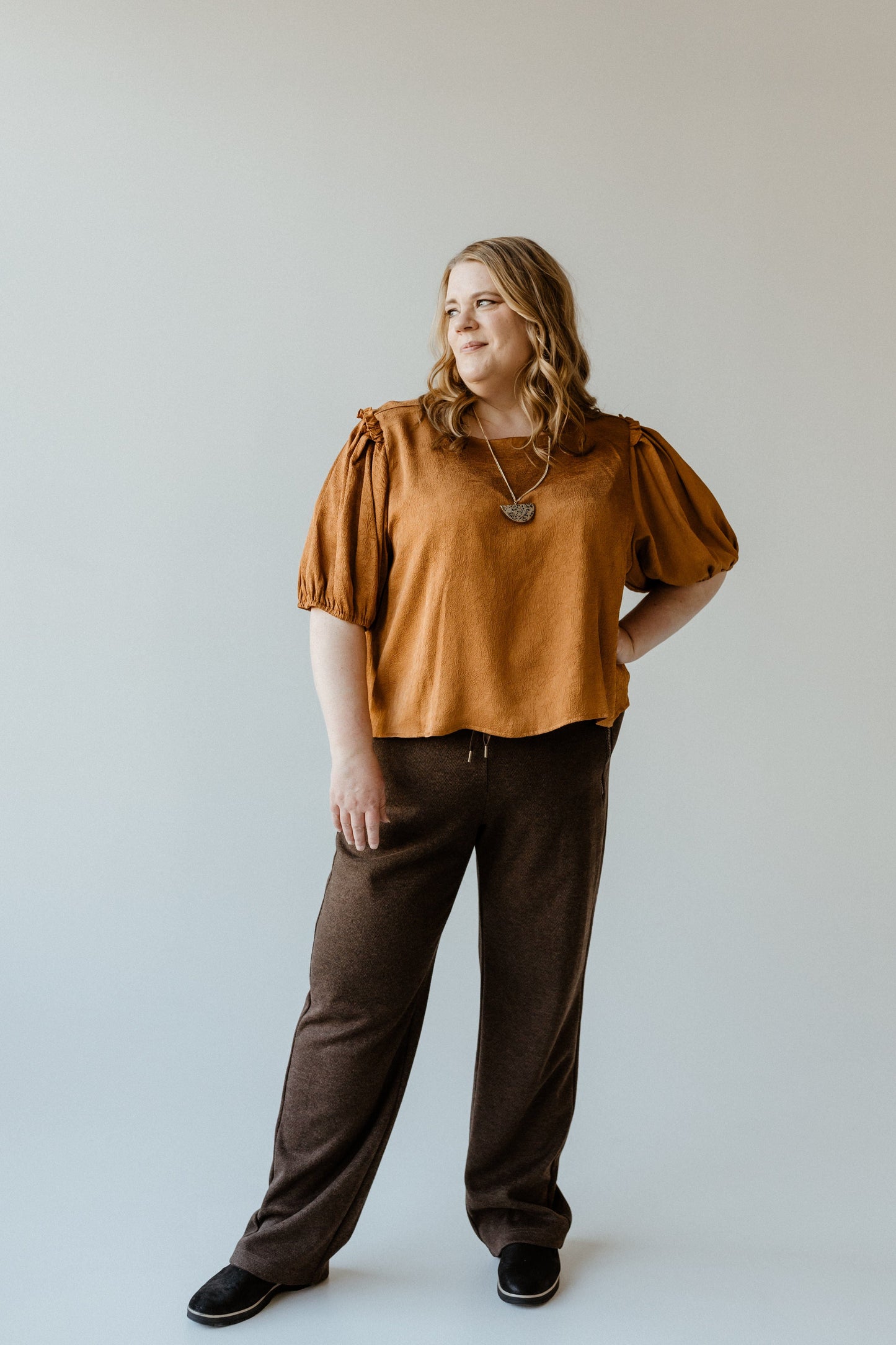 A person stands against a plain background, wearing a brown top and loose high-waist, wide-leg trousers paired with black shoes, looking off to the side.