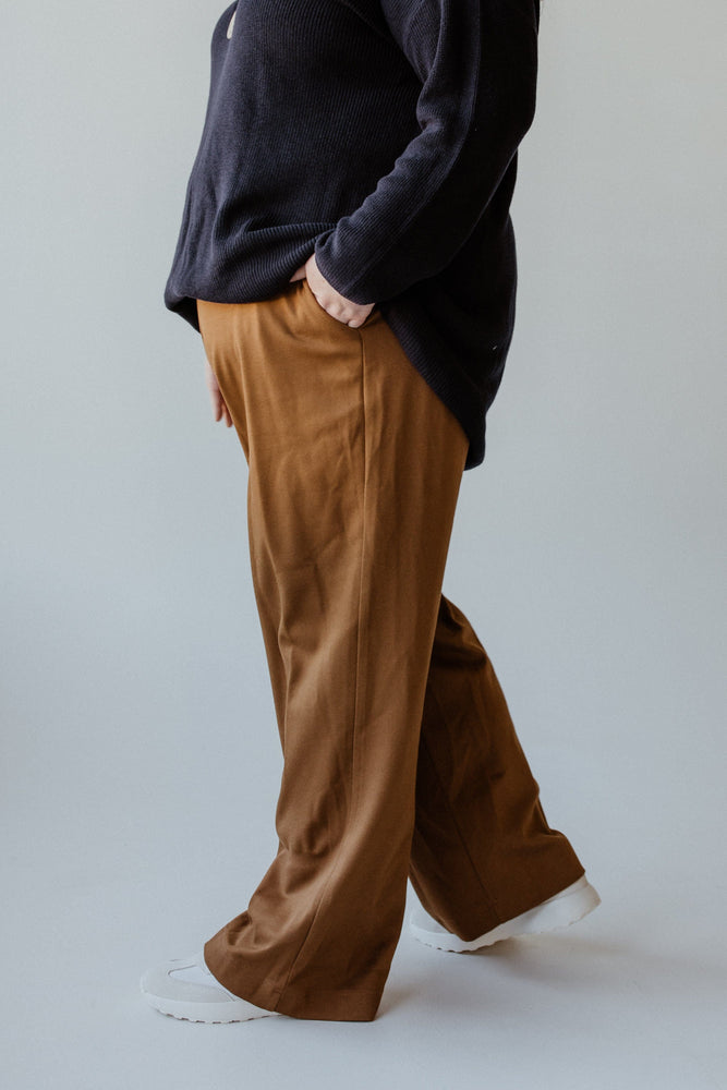 A person wearing the Yesta WIDE LEG TREGGING WITH POCKETS IN REGULAR in brown, featuring a comfortable pull-on waistband, pairs it with a black long-sleeve shirt and white shoes while standing with one hand in their pocket against a plain background.