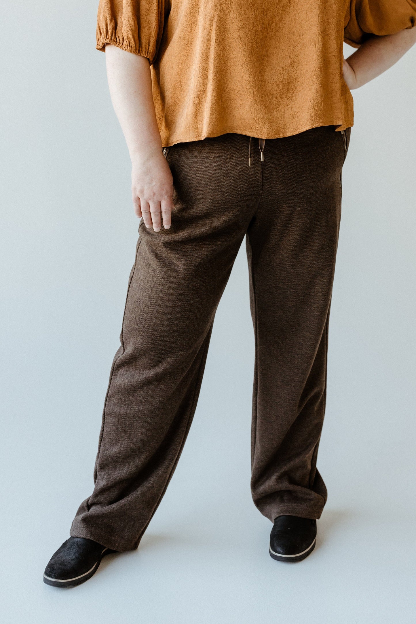 A person in a brown top and high waist, wide-leg trousers steps confidently, black shoes grounding the look against a light gray background.