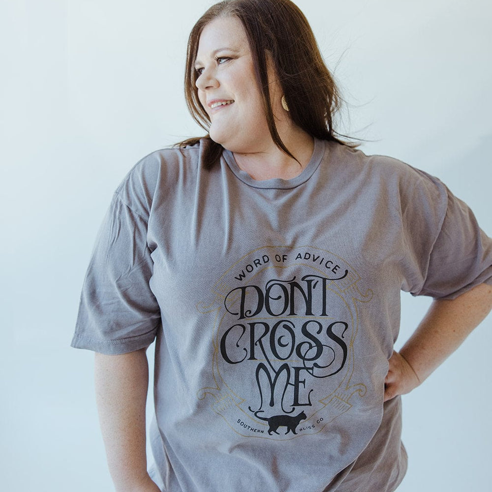 A person with long hair wears the Love Marlow "WORD OF ADVICE DON'T CROSS ME" graphic tee in gray, set against a light-colored background while smiling and looking to the side. The T-shirt's vintage wash enhances their laid-back style with a hint of retro flair.