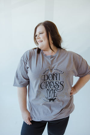 A person with long hair wears the Love Marlow "WORD OF ADVICE DON'T CROSS ME" graphic tee in gray, set against a light-colored background while smiling and looking to the side. The T-shirt's vintage wash enhances their laid-back style with a hint of retro flair.