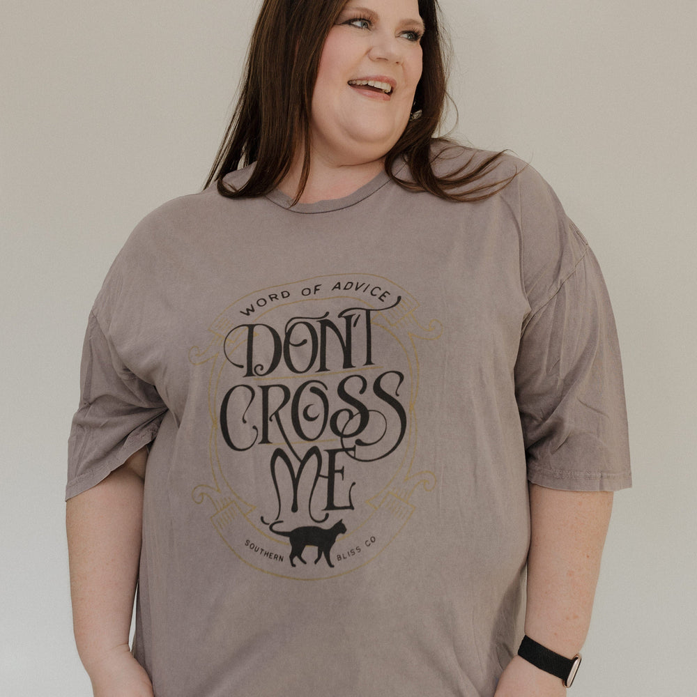 "WORD OF ADVICE DON'T CROSS ME" GRAPHIC TEE A person with long hair wears the Love Marlow "WORD OF ADVICE DON'T CROSS ME" graphic tee in gray, set against a light-colored background while smiling and looking to the side. The T-shirt's vintage wash enhances their laid-back style with a hint of retro flair.