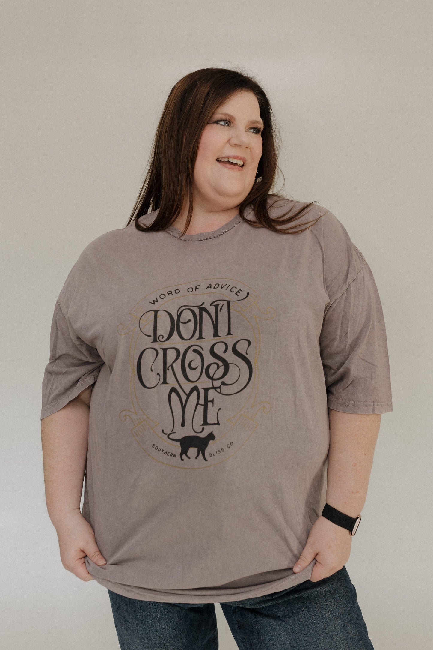 "WORD OF ADVICE DON'T CROSS ME" GRAPHIC TEE A person with long hair wears the Love Marlow "WORD OF ADVICE DON'T CROSS ME" graphic tee in gray, set against a light-colored background while smiling and looking to the side. The T-shirt's vintage wash enhances their laid-back style with a hint of retro flair.