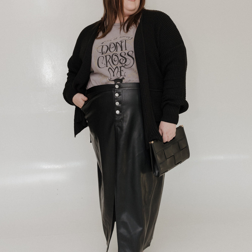 "WORD OF ADVICE DON'T CROSS ME" GRAPHIC TEE A person with long hair wears the Love Marlow "WORD OF ADVICE DON'T CROSS ME" graphic tee in gray, set against a light-colored background while smiling and looking to the side. The T-shirt's vintage wash enhances their laid-back style with a hint of retro flair. Pleather skirt and purse.