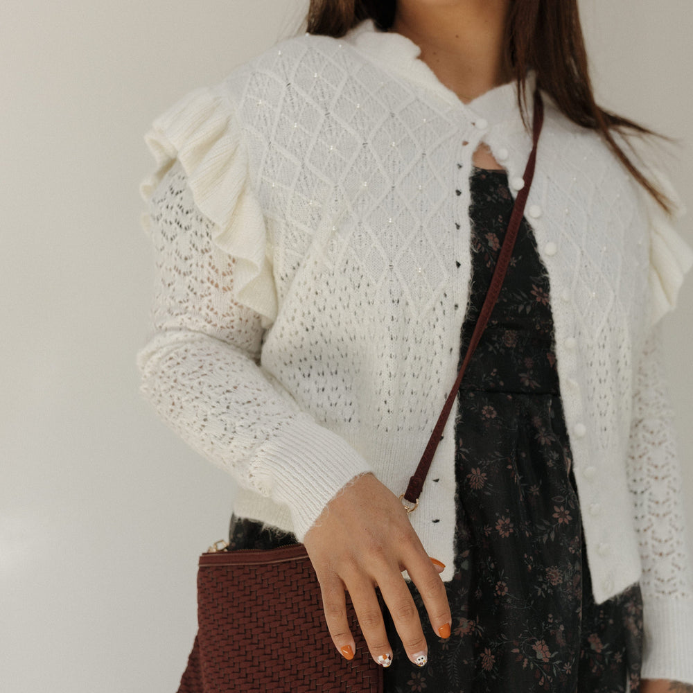 Woman wearing a white textured cardigan over a dark floral dress, holding a brown woven handbag.