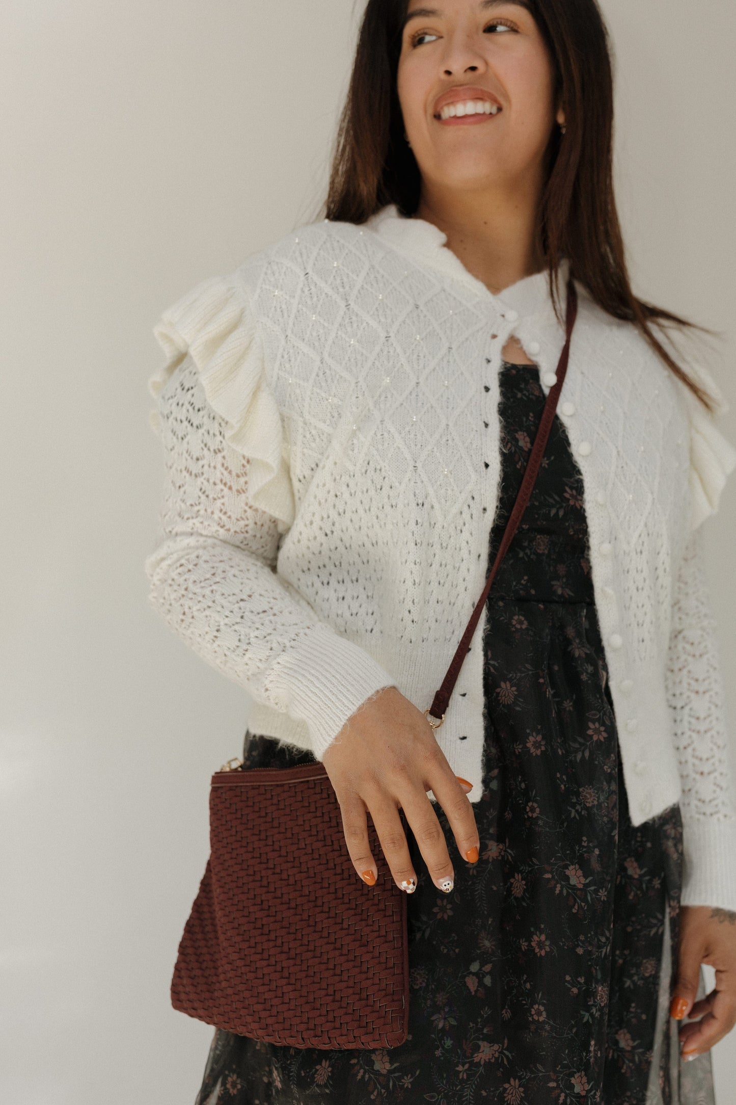 Woman wearing a white textured cardigan over a dark floral dress, holding a brown woven handbag.