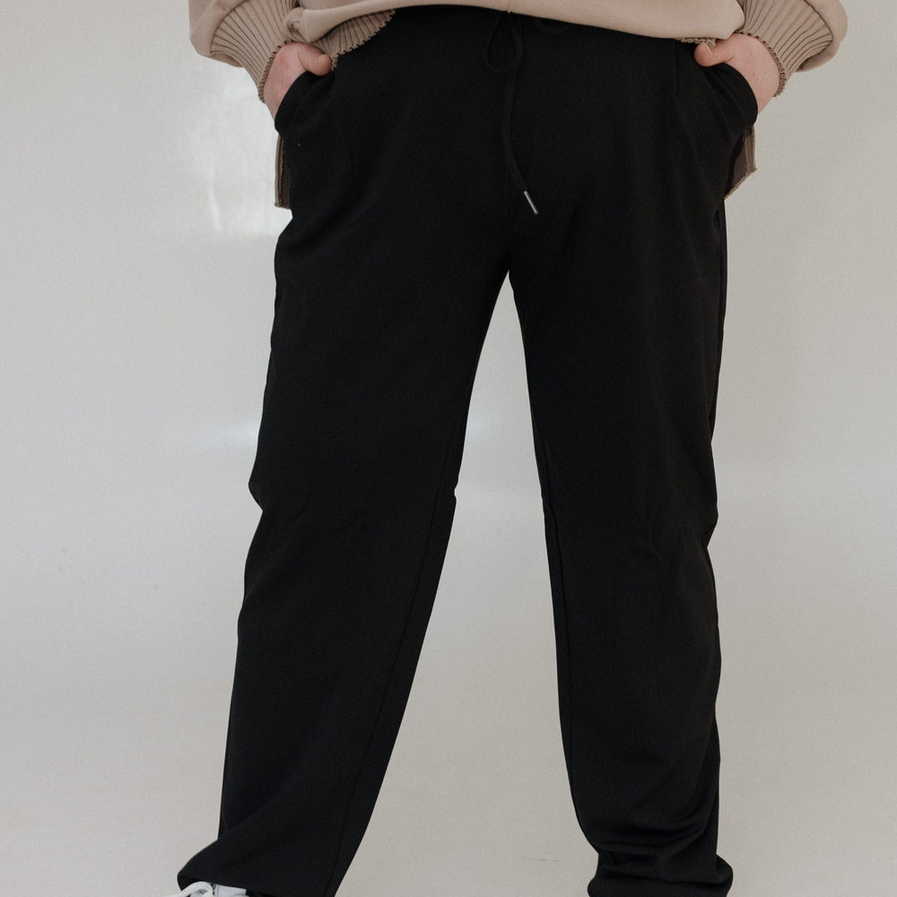 Person wearing a beige sweatshirt and black pants with white sneakers on a plain background