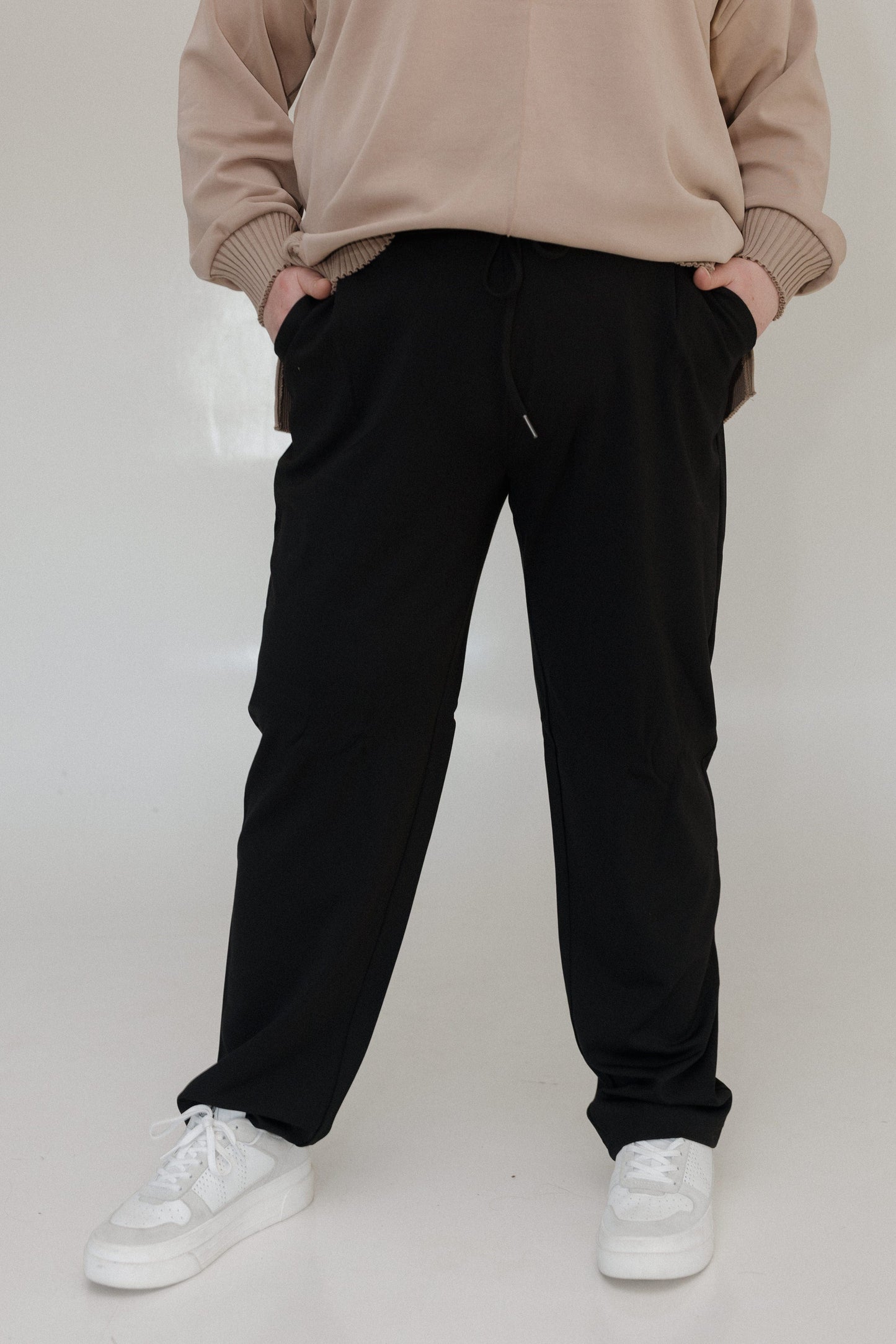 Person wearing a beige sweatshirt and black pants with white sneakers on a plain background
