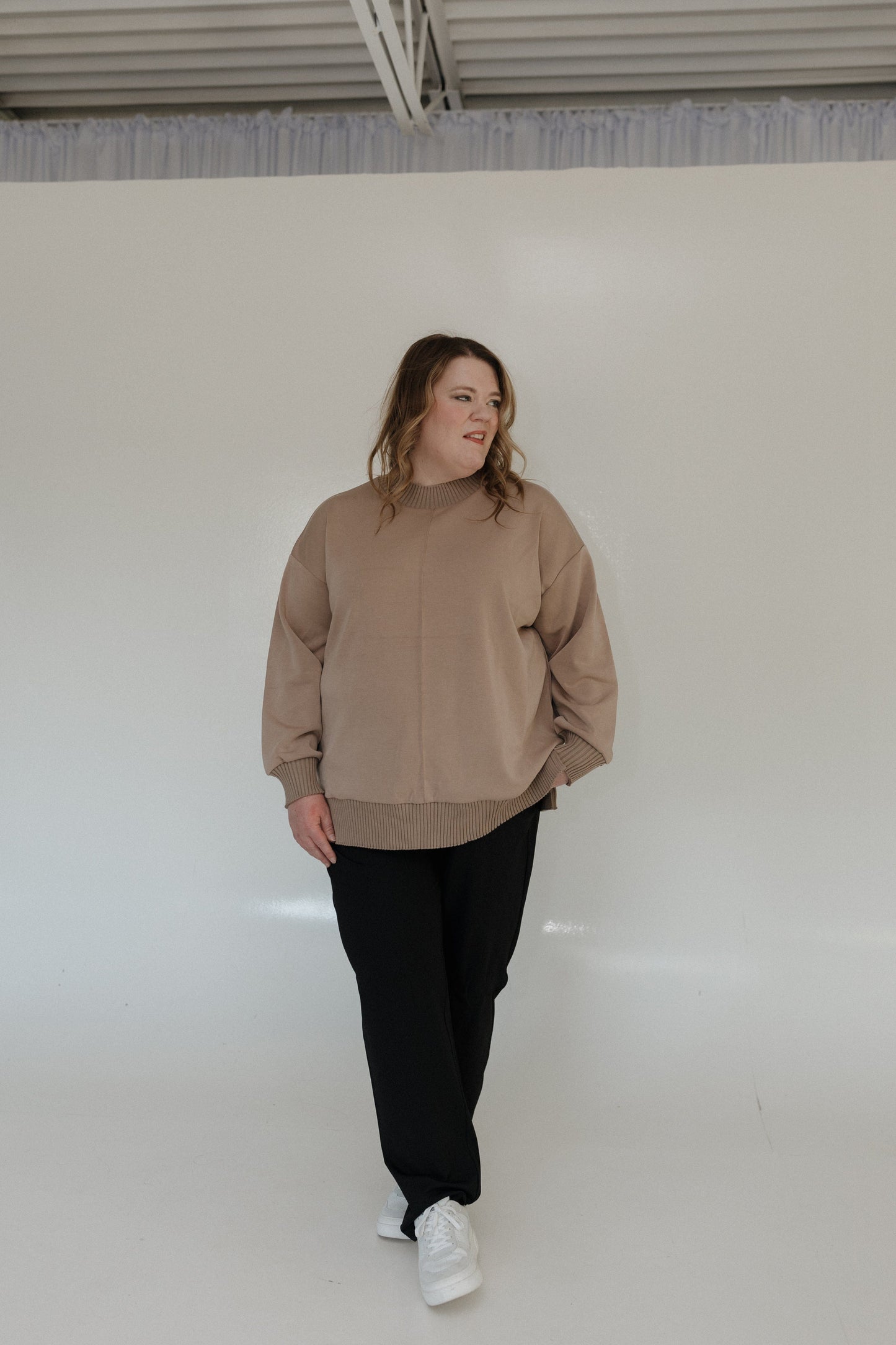 Person wearing a beige sweater and black pants standing against a white wall.