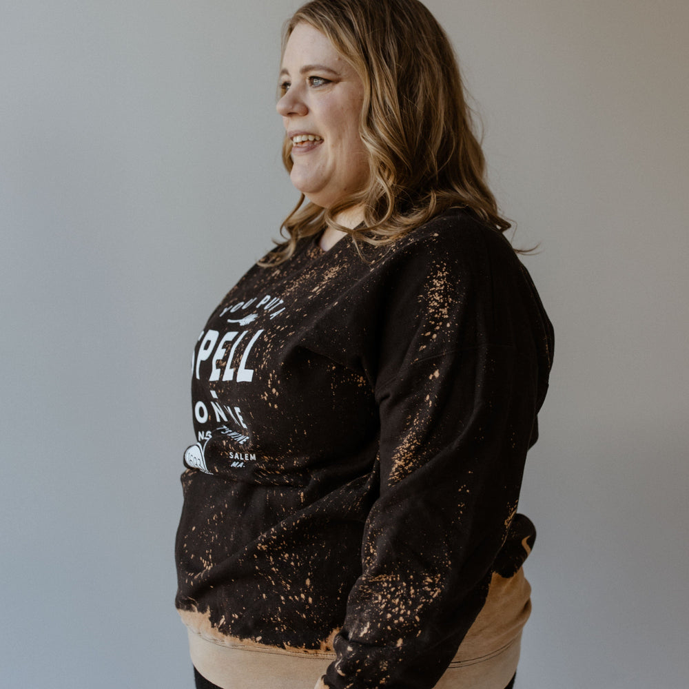 A woman with wavy hair is wearing a black and orange "YOU PUT A SPELL ON ME NOW LET'S WINE" graphic sweatshirt by Love Marlow, showcasing a striking splatter bleach design.