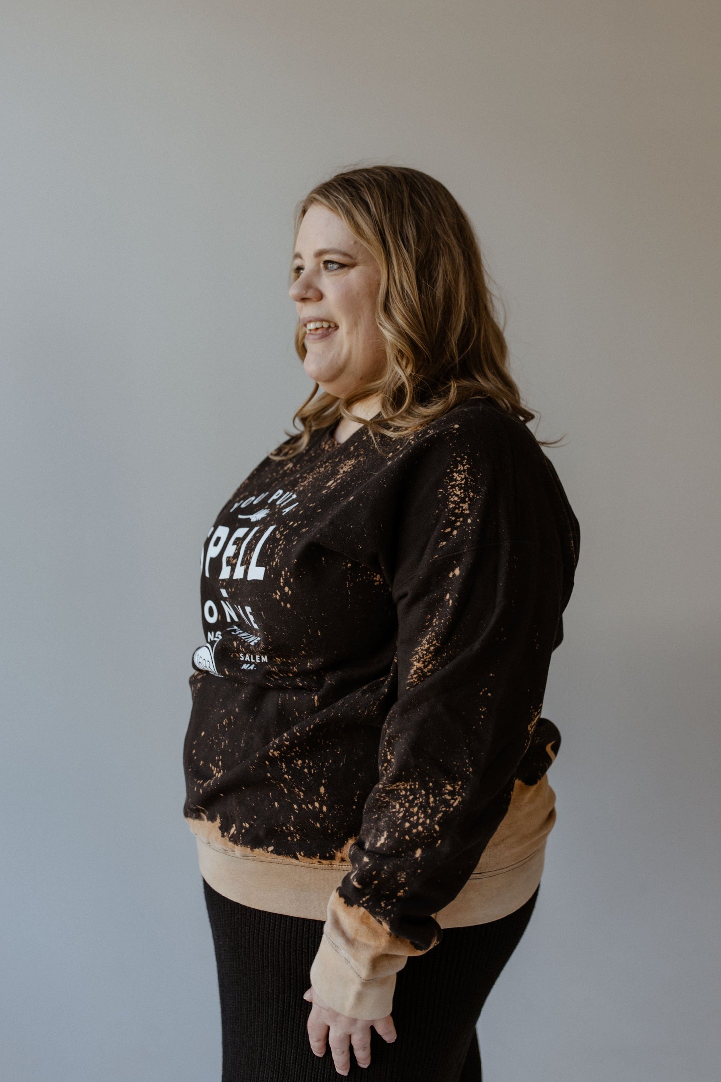 A woman with wavy hair is wearing a black and orange "YOU PUT A SPELL ON ME NOW LET'S WINE" graphic sweatshirt by Love Marlow, showcasing a striking splatter bleach design.