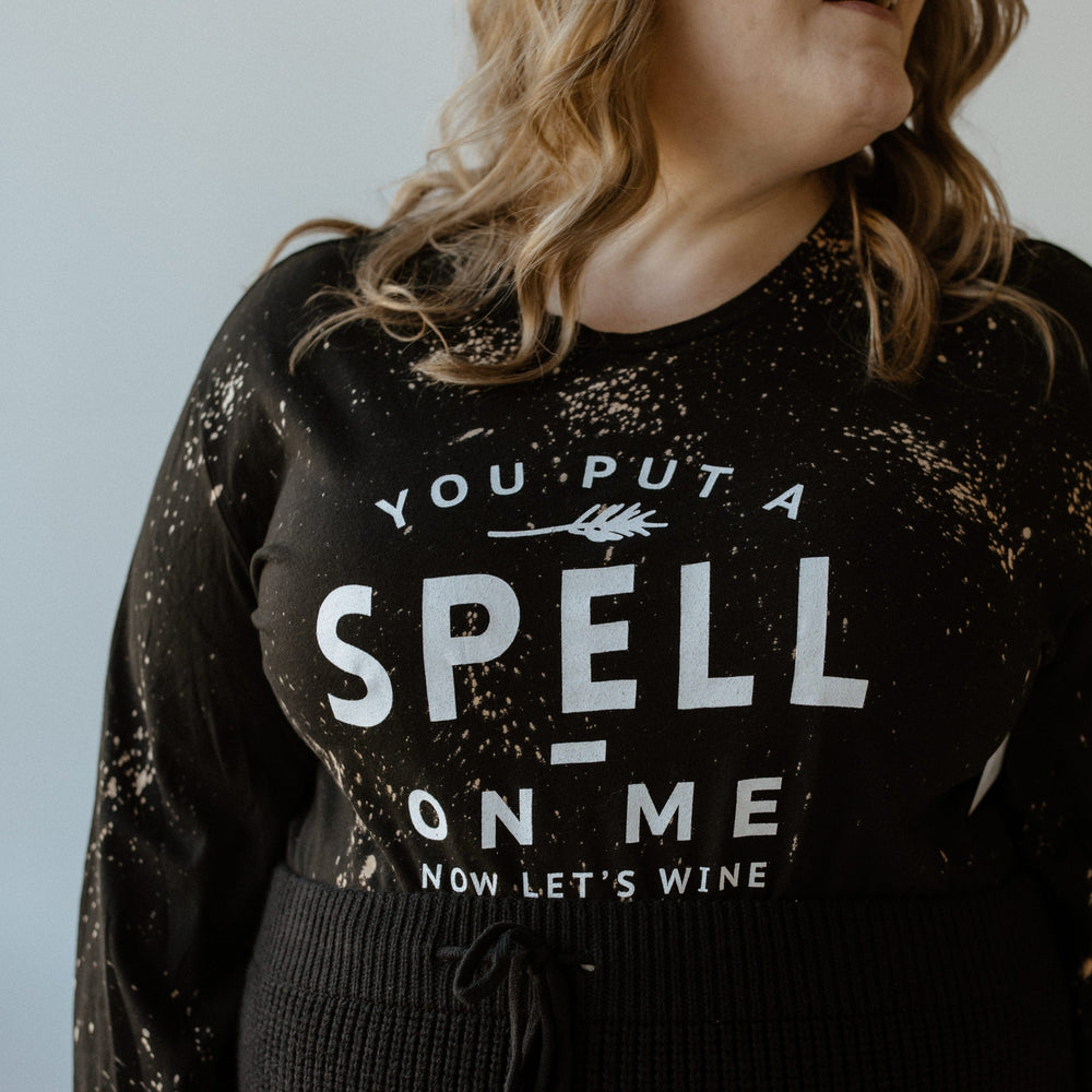 The person is smiling and looking to the side, wearing a black long sleeve graphic tee by Love Marlow featuring the text "YOU PUT A SPELL ON ME NOW LET'S WINE" in white.