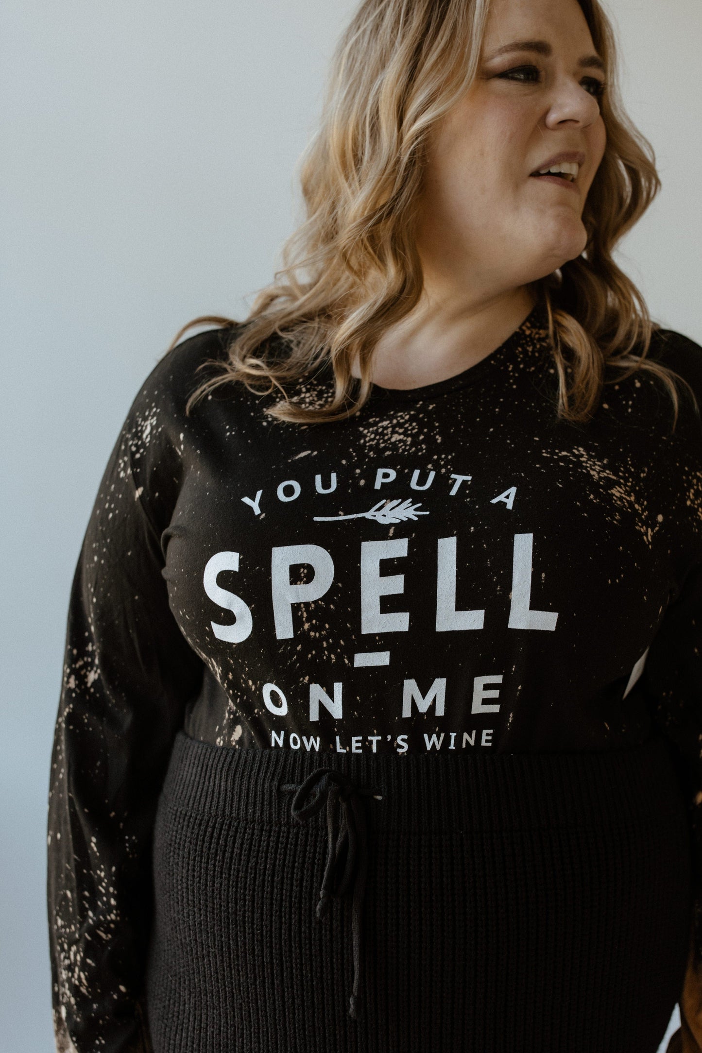 The person is smiling and looking to the side, wearing a black long sleeve graphic tee by Love Marlow featuring the text "YOU PUT A SPELL ON ME NOW LET'S WINE" in white.