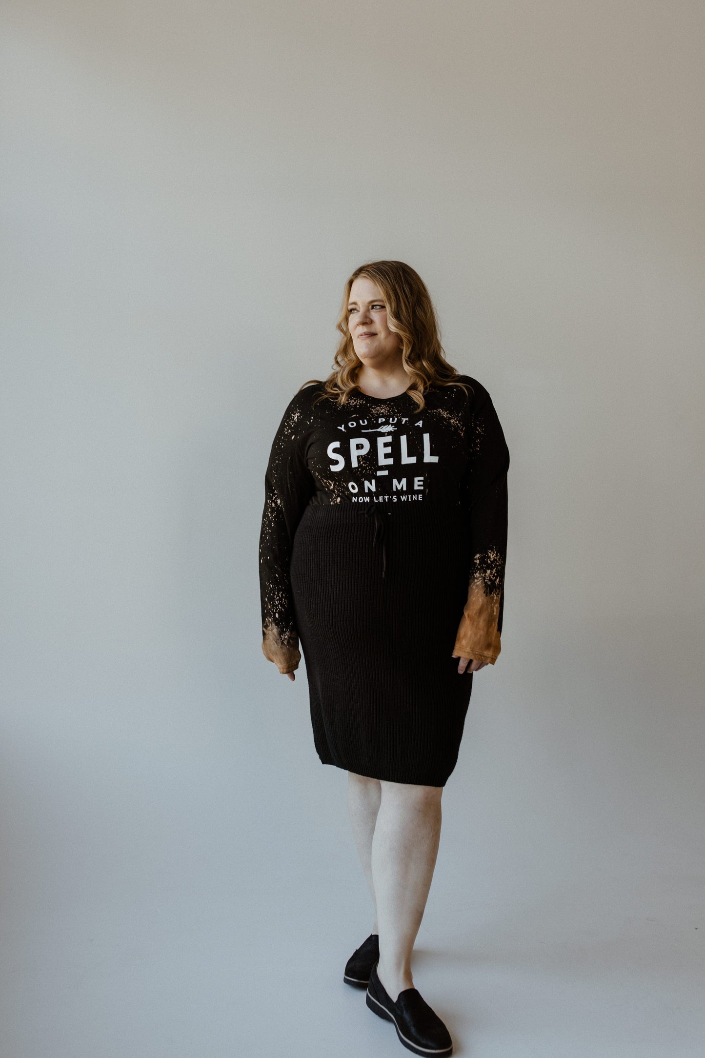 A person dressed in Love Marlow's long sleeve graphic tee, featuring the playful text "You Put a Spell on Me Now Let's Wine," stands against a plain backdrop, exuding Halloween vibes.