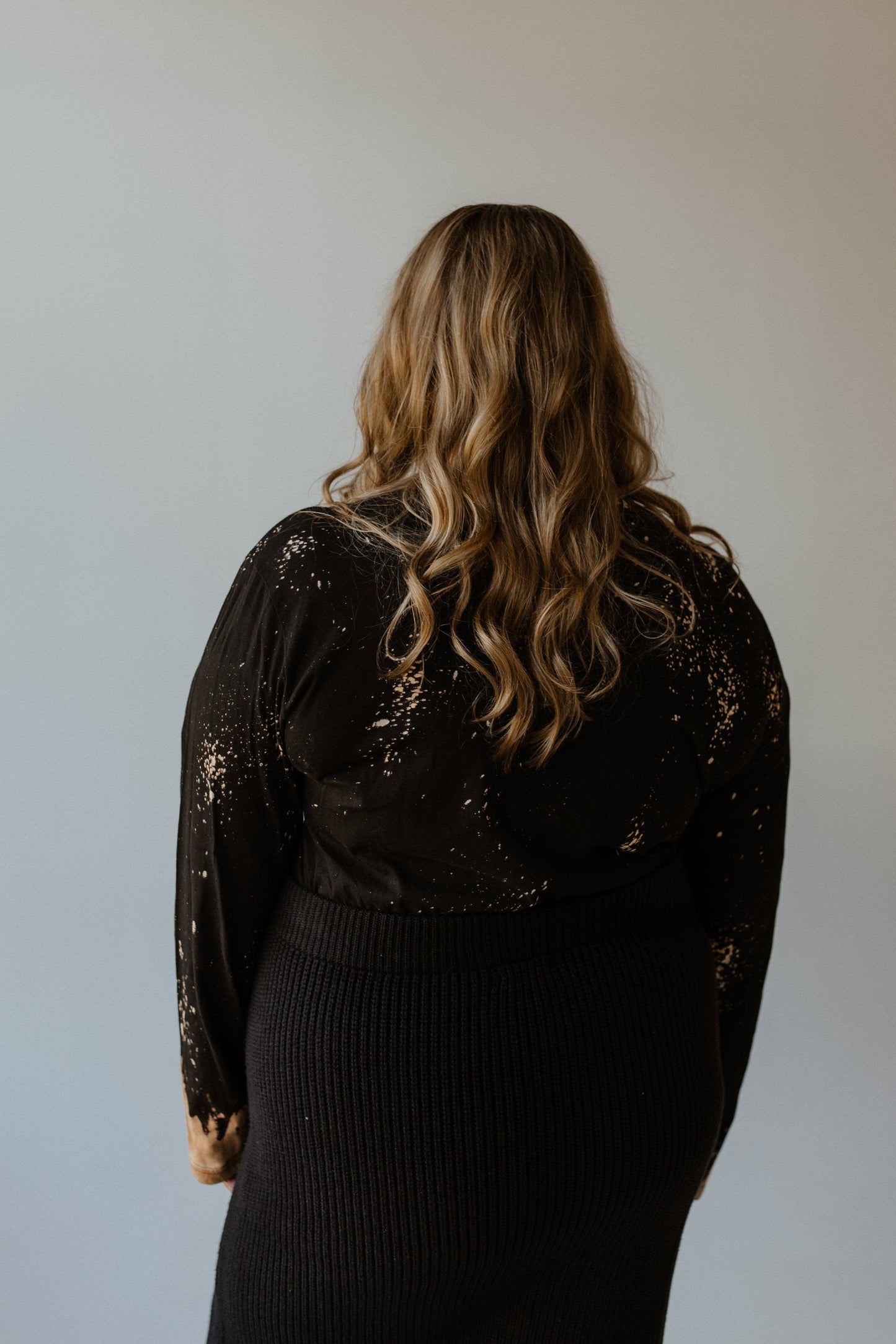 The individual with long wavy hair is seen from behind against a neutral background, wearing the 