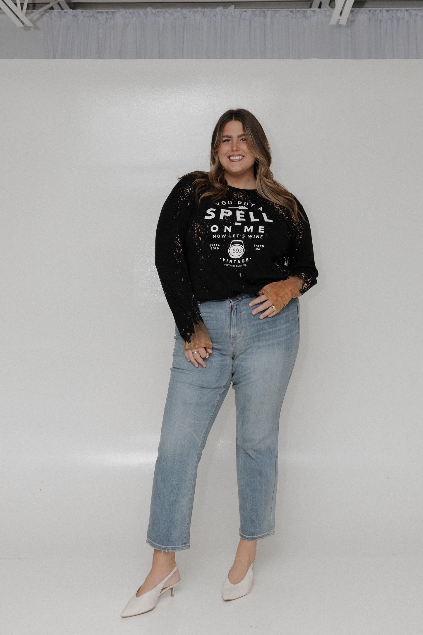 "YOU PUT A SPELL ON ME NOW LET'S WINE" LONG SLEEVE GRAPHIC TEE Woman wearing a black sweatshirt with text and sheer sleeves against a white background at Love Marlow