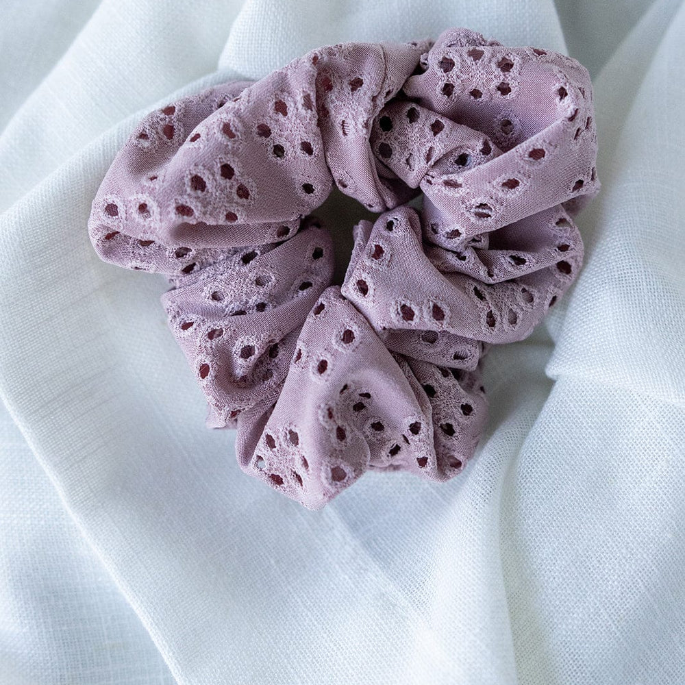 A mauve eyelet lace scrunchie delicately rests on a white fabric background.