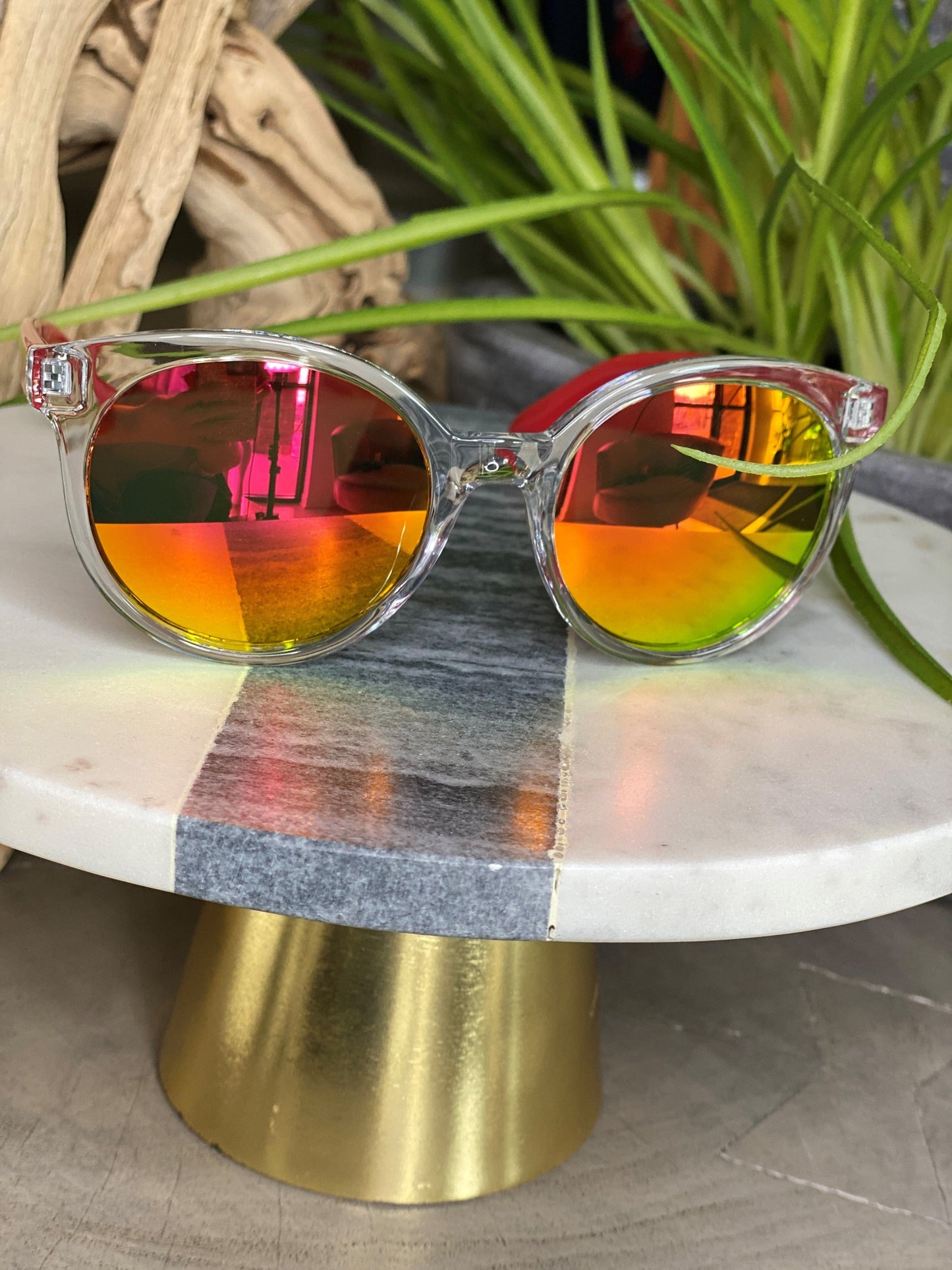 The Love Marlow BARBADOS SUNGLASSES IN RED, featuring clear frames and reflective red and yellow lenses that offer UVA/UVB protection, are elegantly displayed on a marble and gold stand against a backdrop of lush green plants.