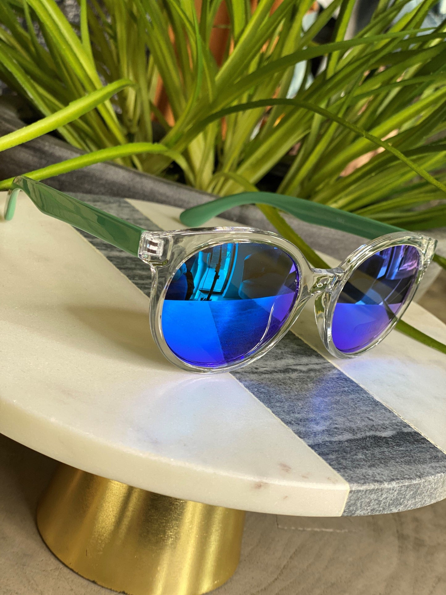 A pair of Love Marlow BARBADOS SUNGLASSES IN BLUE, offering UVA/UVB protection, rests on a round, white and gray marble surface with green plants in the background.