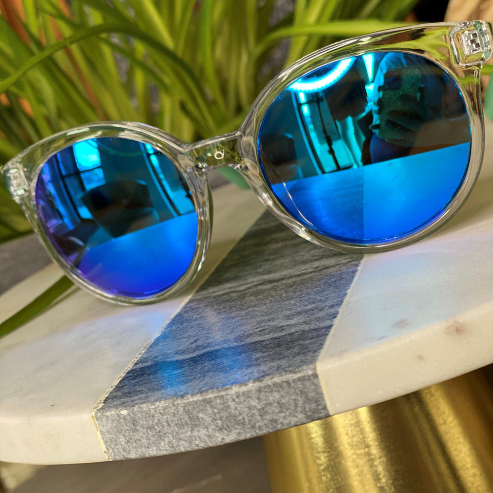 BARBADOS SUNGLASSES IN BLUE by Love Marlow, offering UVA/UVB protection, are placed on a marble table with a plant in the background.