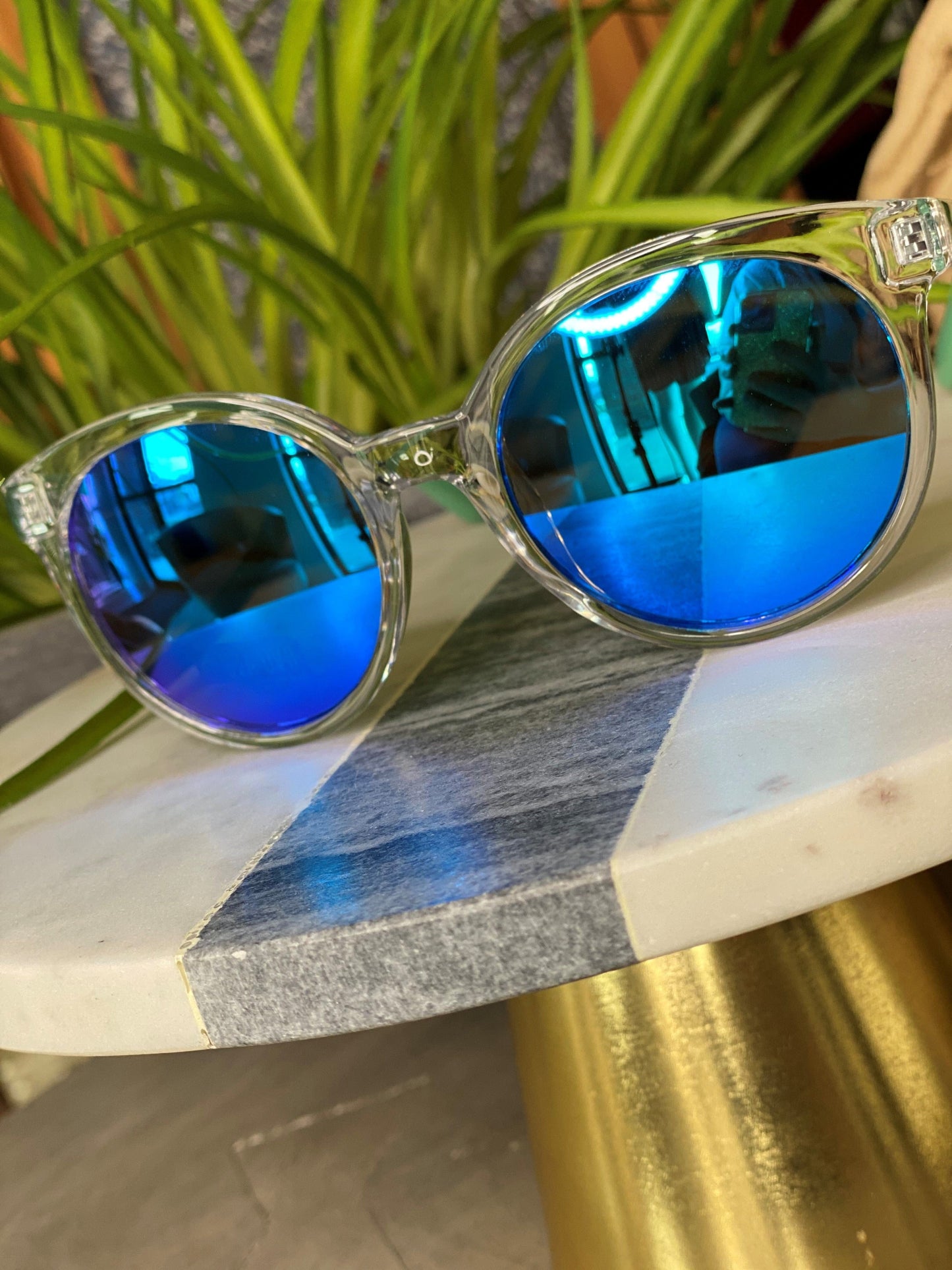 BARBADOS SUNGLASSES IN BLUE by Love Marlow, offering UVA/UVB protection, are placed on a marble table with a plant in the background.