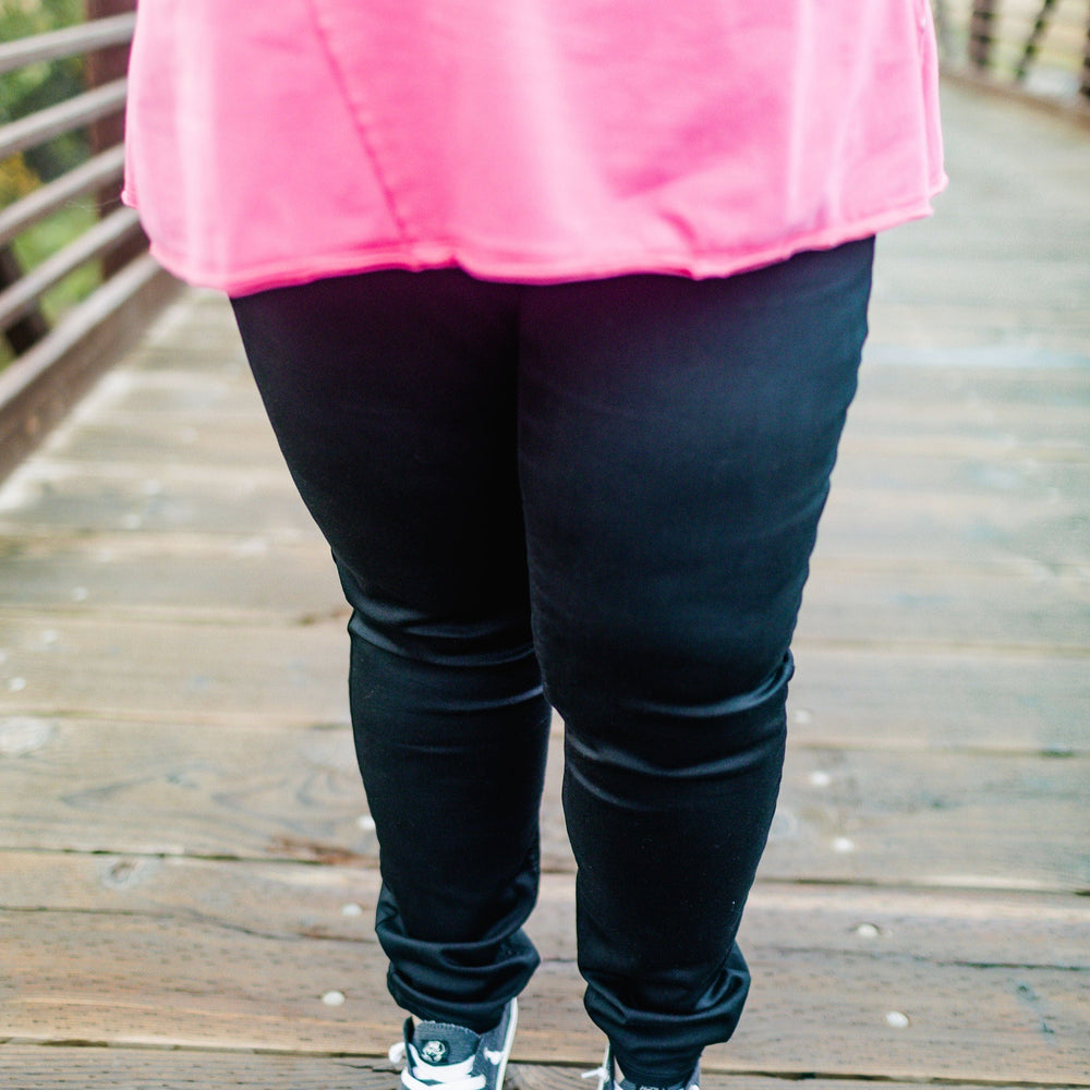 A person wearing a pink shirt and high-rise jeggings in a charcoal color, paired with black sneakers with white laces, stands confidently on a wooden bridge.