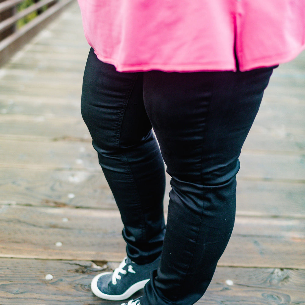 A person stands on a wooden bridge, wearing a pink shirt and black sneakers. Their outfit is completed with charcoal-colored high-rise jeggings made of stretchy fabric, creating a stylish and comfortable look.