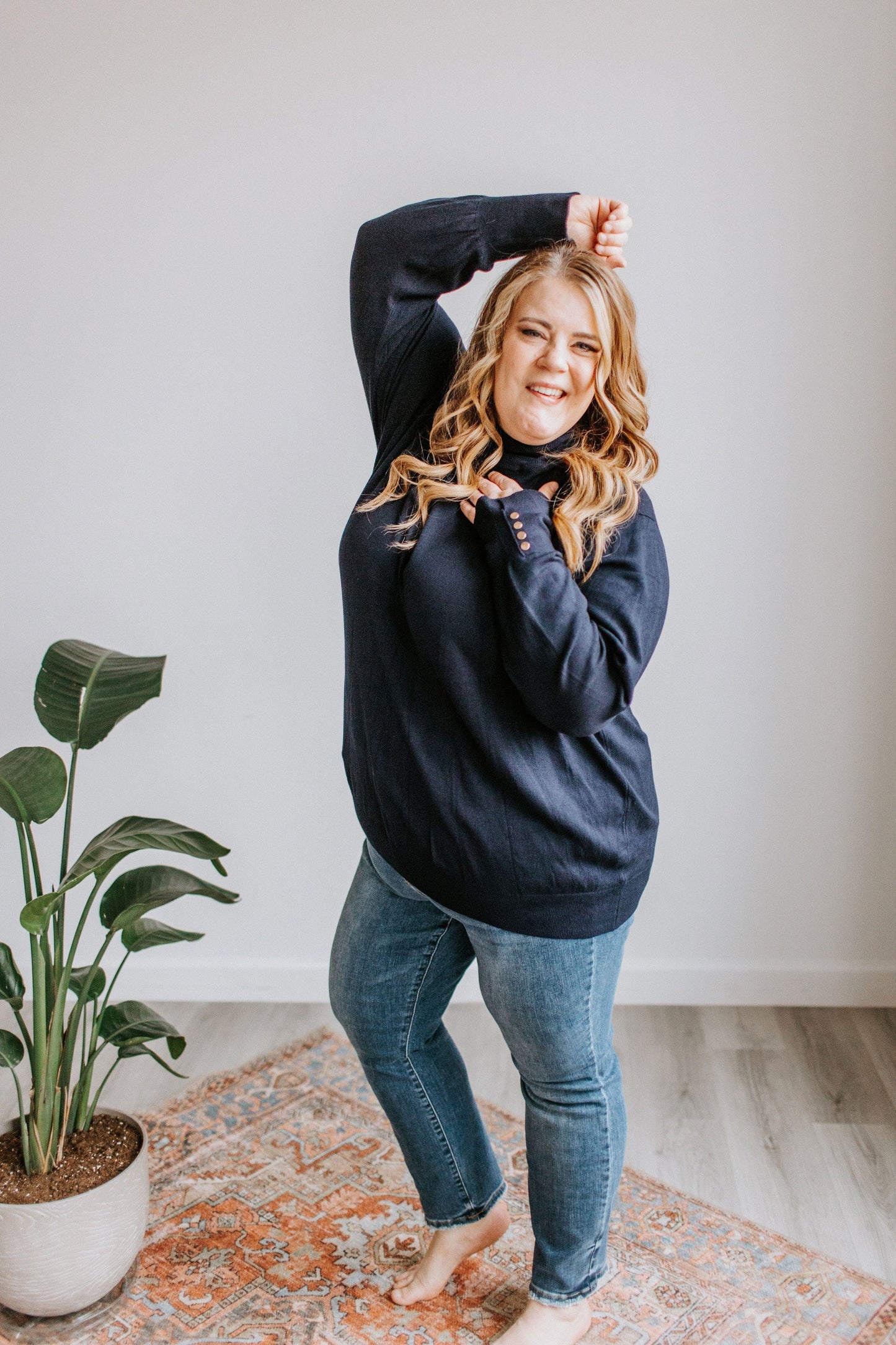 A woman with long hair smiles and poses playfully, standing on a patterned rug beside a potted plant. She wears a luxurious navy turtleneck sweater and blue jeans.