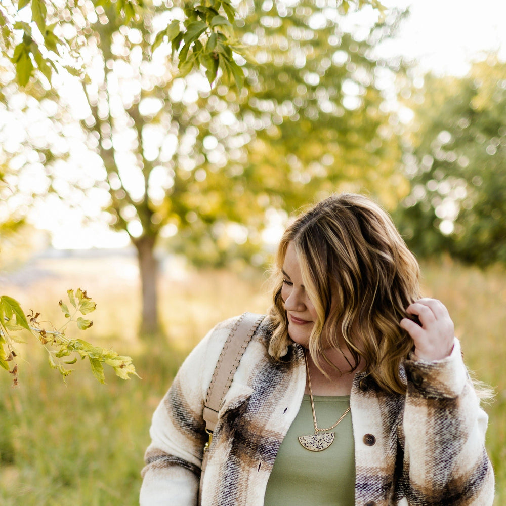 A woman with shoulder-length hair in a plaid jacket stands outdoors, brushing her hair back. A jasper pendant gleams against her outfit as she basks in the grassy area, with trees and sunlight filtering through.