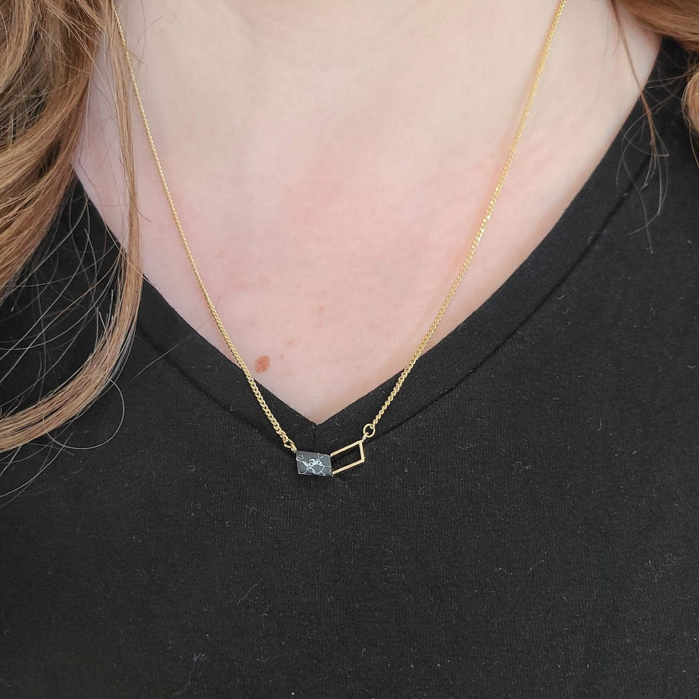 A Corinne Chevron Necklace featuring a dainty gold chain and a black marbled chevron pendant, gracefully complements the geometric charm as it's worn over a sleek black top.