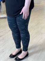 A person wearing Spanx© LOOK AT ME NOW SEAMLESS LEGGING in a black camouflage design, featuring a double-layer waistband for shaping support, stands on a light wood floor, paired elegantly with black flats.