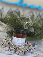 A brown glass jar candle with a black lid, labeled "Love Marlow HAND POURED SOY WAX CANDLES," made from soy wax and premium fragrance oils, is placed on a cream-colored knit blanket.