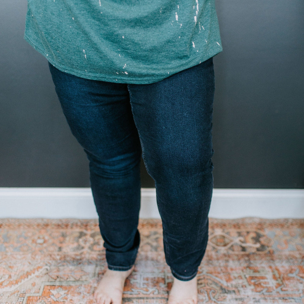 A person in a green shirt and stretchy jeggings stands barefoot on a patterned rug.