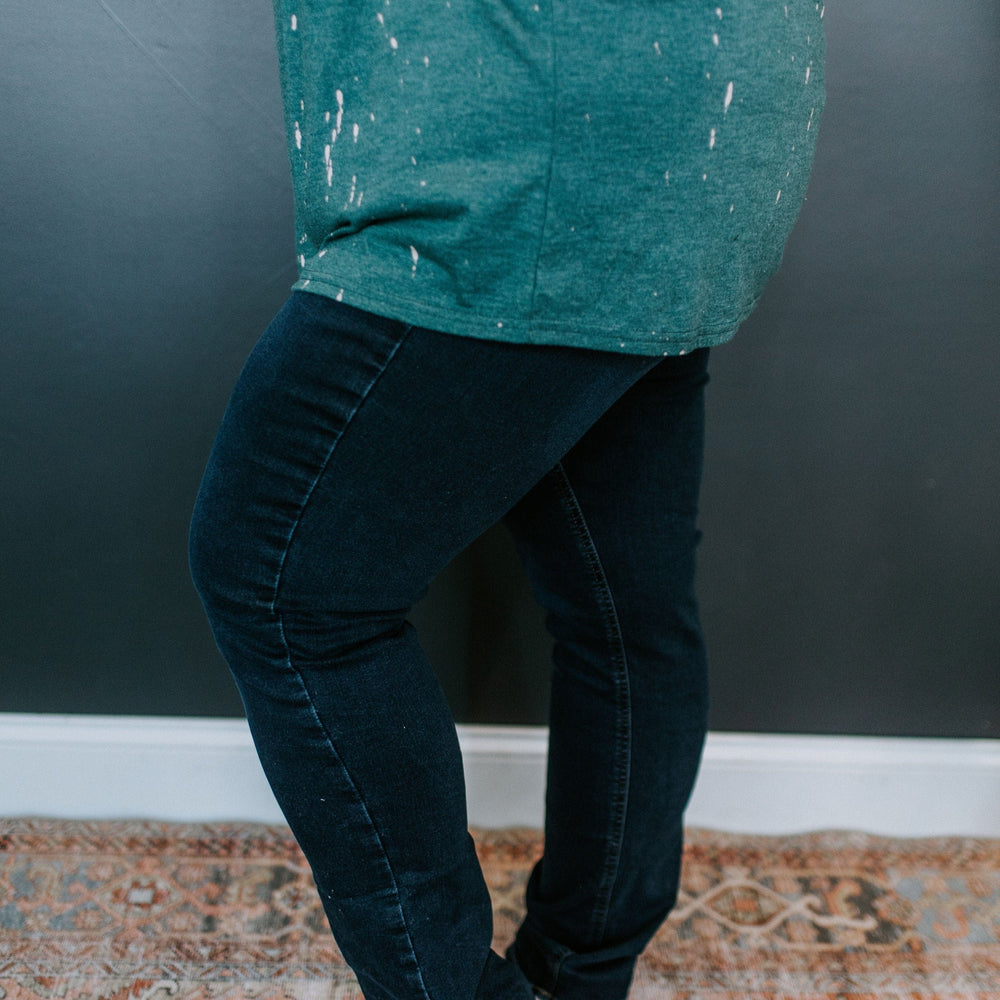 A person stands barefoot on a rug, wearing a paint-splattered green shirt and dark high-rise jeggings with one hand on their hip.