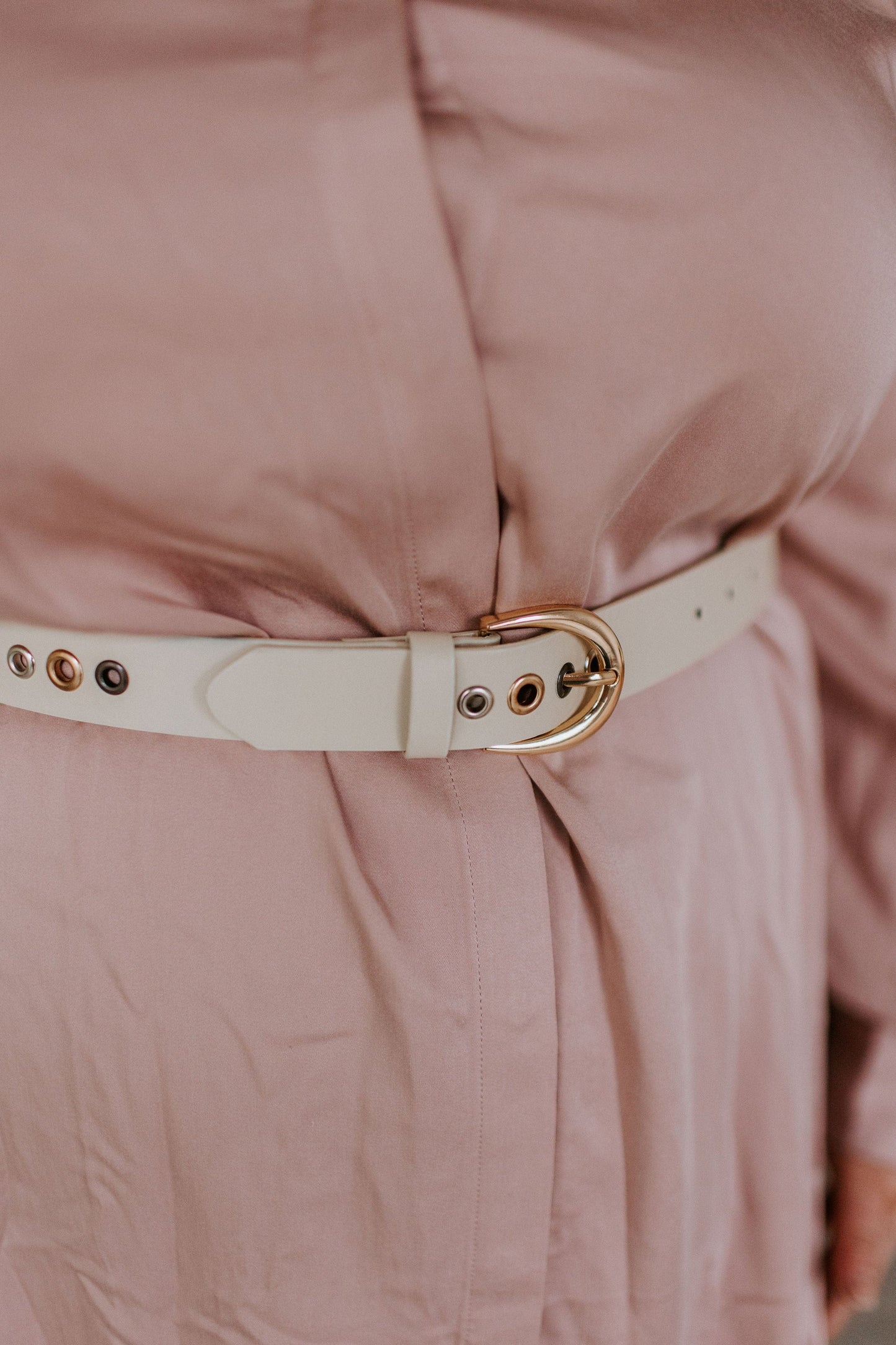 Close-up of a waist-defining white faux leather belt with gold geometric grommets fastened around a light pink garment.