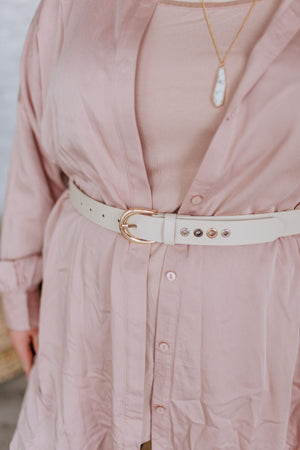 A person wearing a light pink shirt dress cinched with a waist-defining white faux leather belt, accentuated by a necklace featuring a chic white pendant.