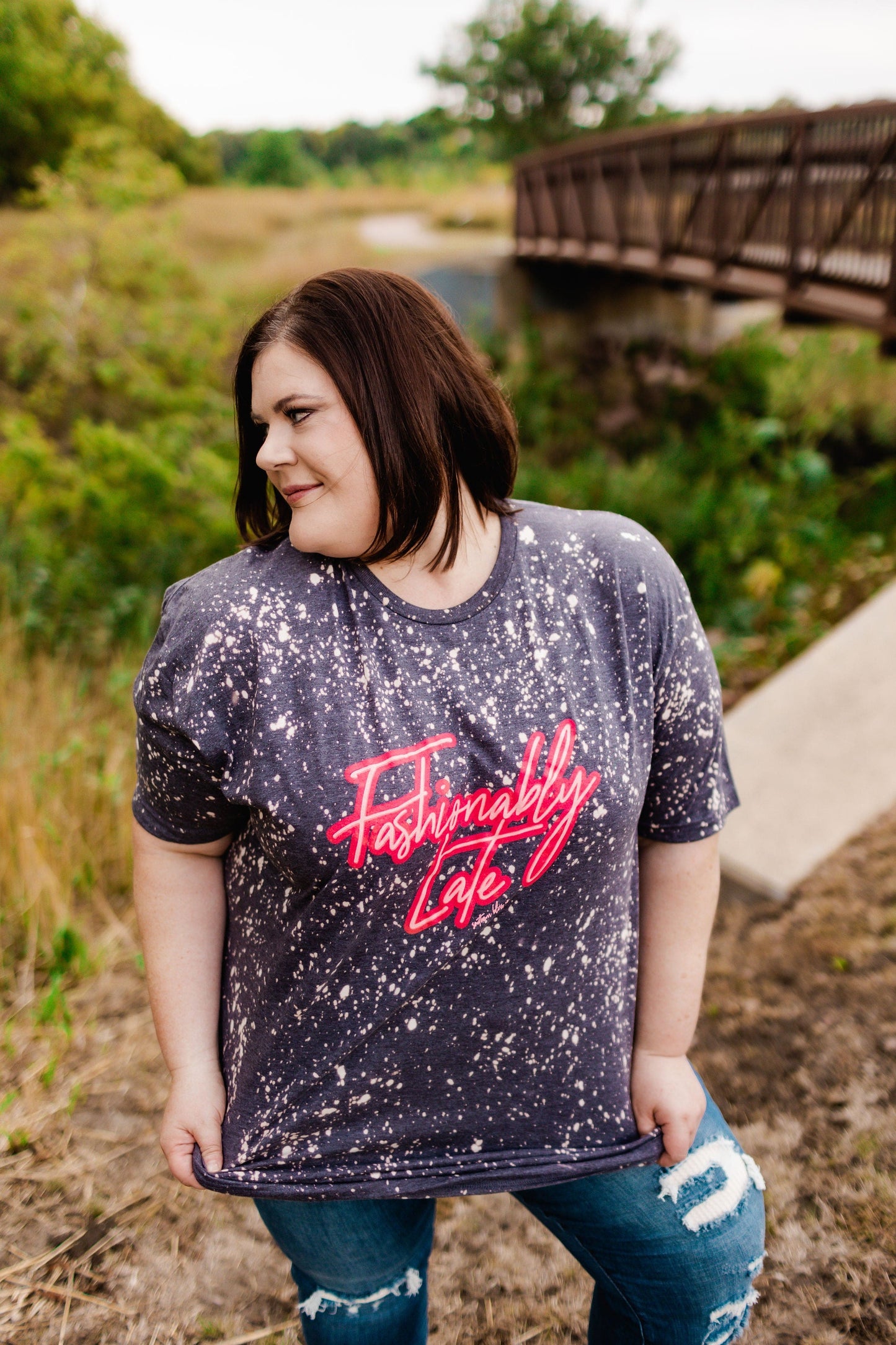 A person wearing a fun warning tee with "Fashionably Late" text stands outdoors near a bridge, looking to the side.