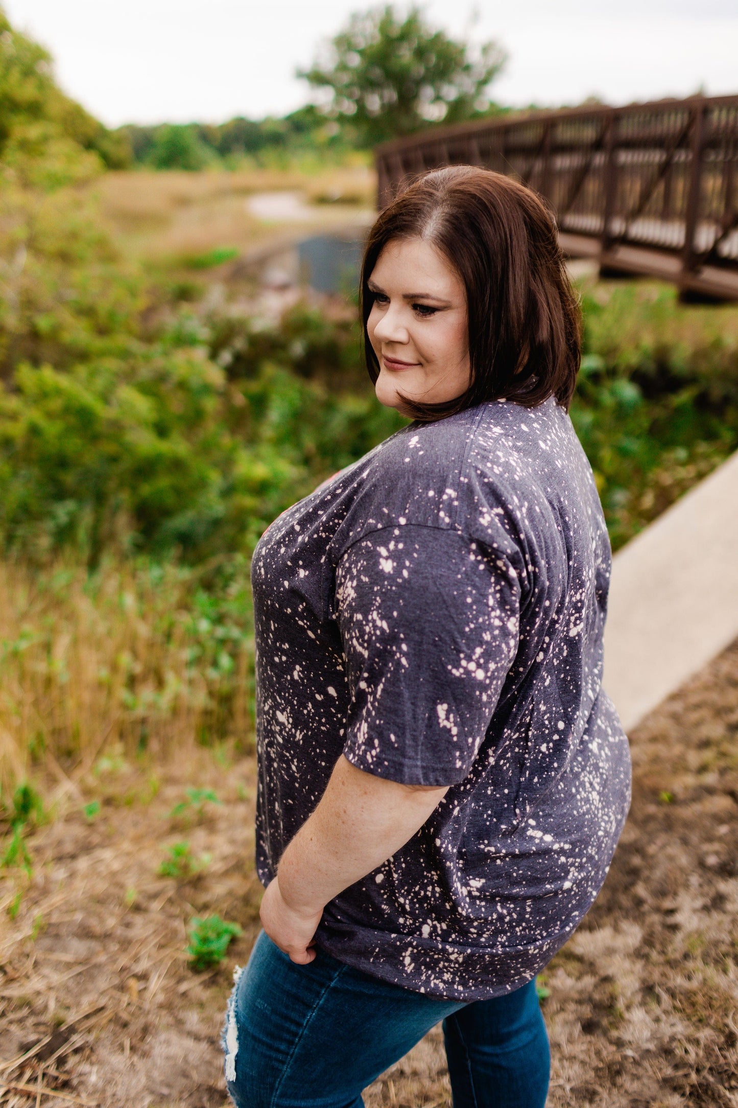 A person in a speckled shirt stands outdoors by a bridge, looking to the side. Lush greenery and a pathway surround the area, adding a vibrant contrast to their heathered mint fabric.