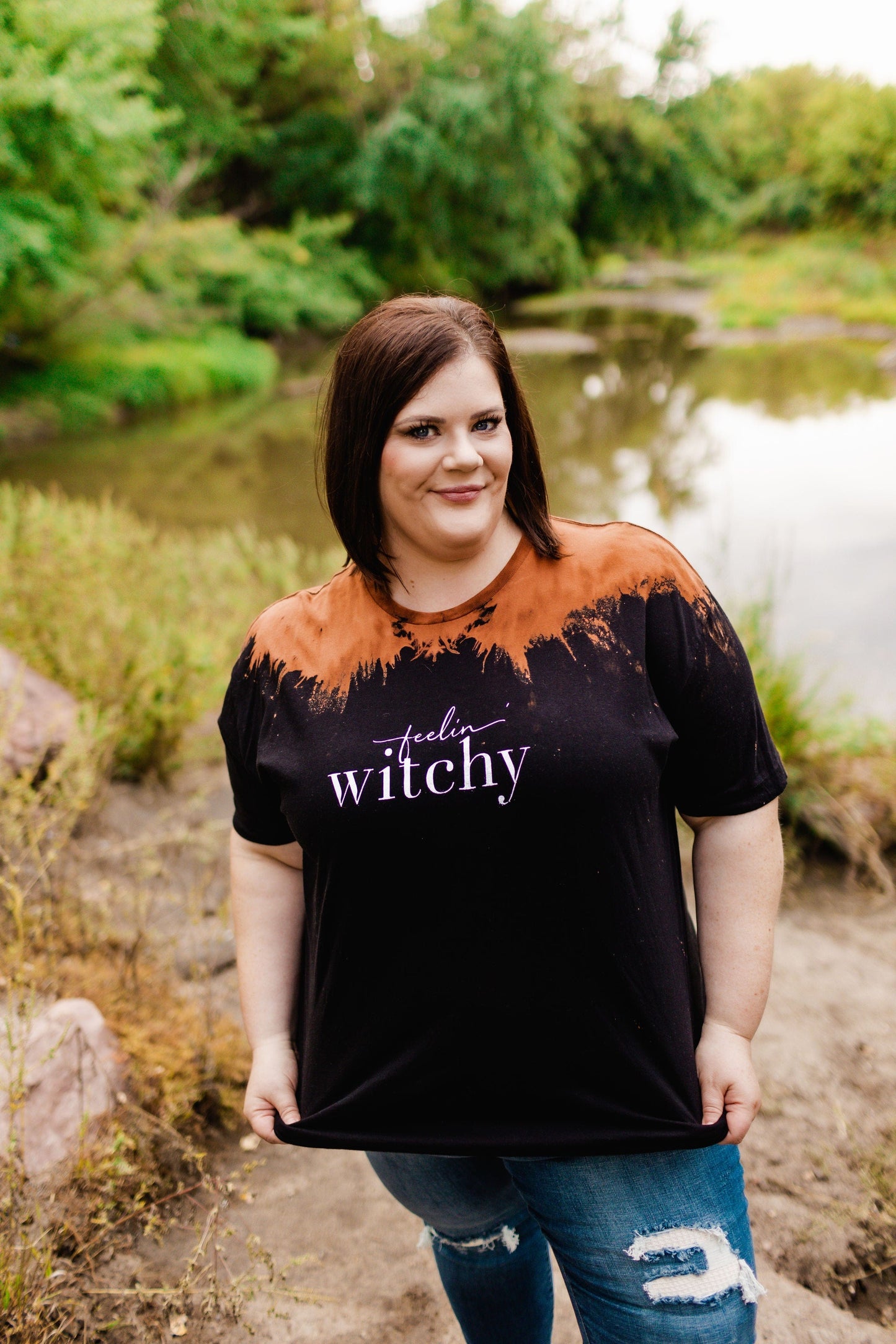 A woman stands by a pond, wearing the "FEELIN' WITCHY" graphic tee by Love Marlow. Smiling and holding the edges of her shirt, she captures the Halloween spirit with an enchanting ombre bleach wash backdrop.