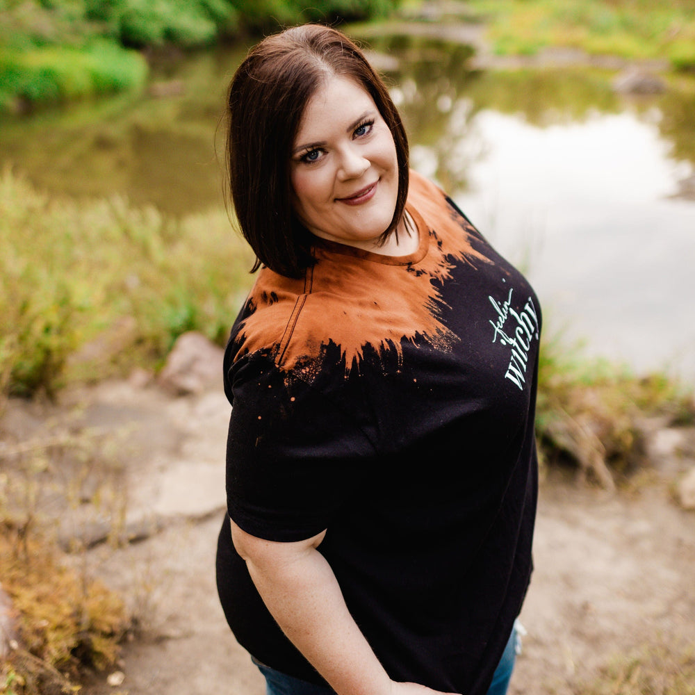 Standing on a rocky path near a pond amidst lush greenery, the person is wearing Love Marlow's "FEELIN' WITCHY" graphic tee in black and orange shades that capture the essence of Halloween, paired with blue jeans.