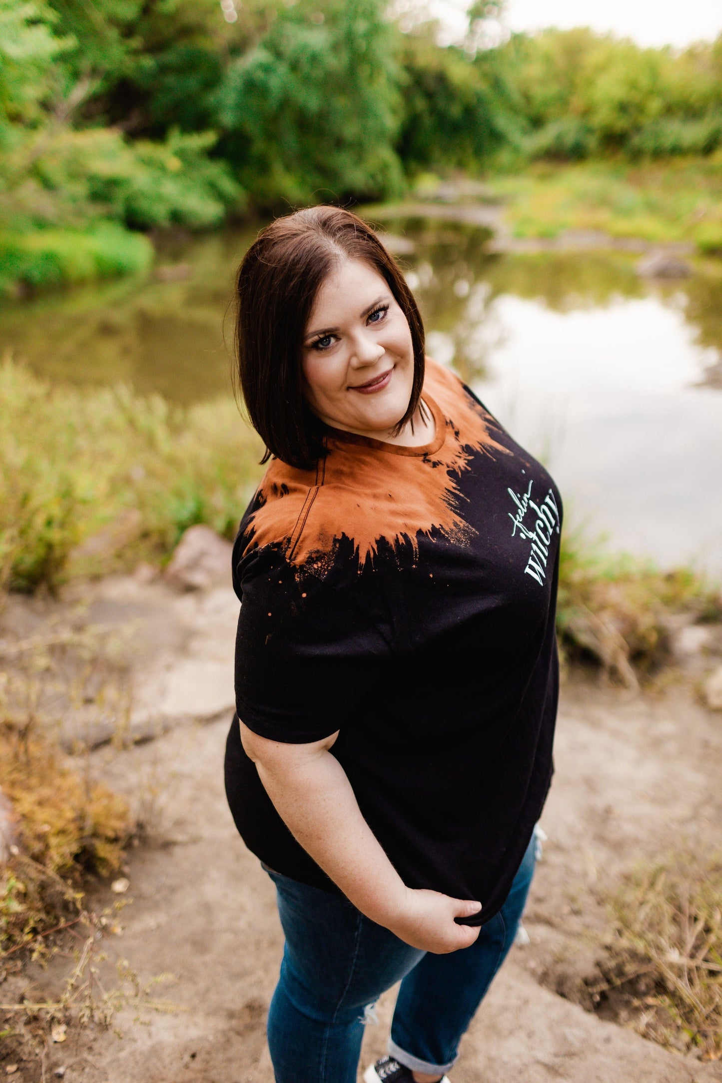 Standing on a rocky path near a pond amidst lush greenery, the person is wearing Love Marlow's "FEELIN' WITCHY" graphic tee in black and orange shades that capture the essence of Halloween, paired with blue jeans.