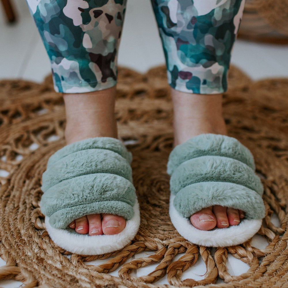 Close-up of feet in green plush slippers with rubber soles on a woven rug. The person is wearing camouflage-patterned pants.