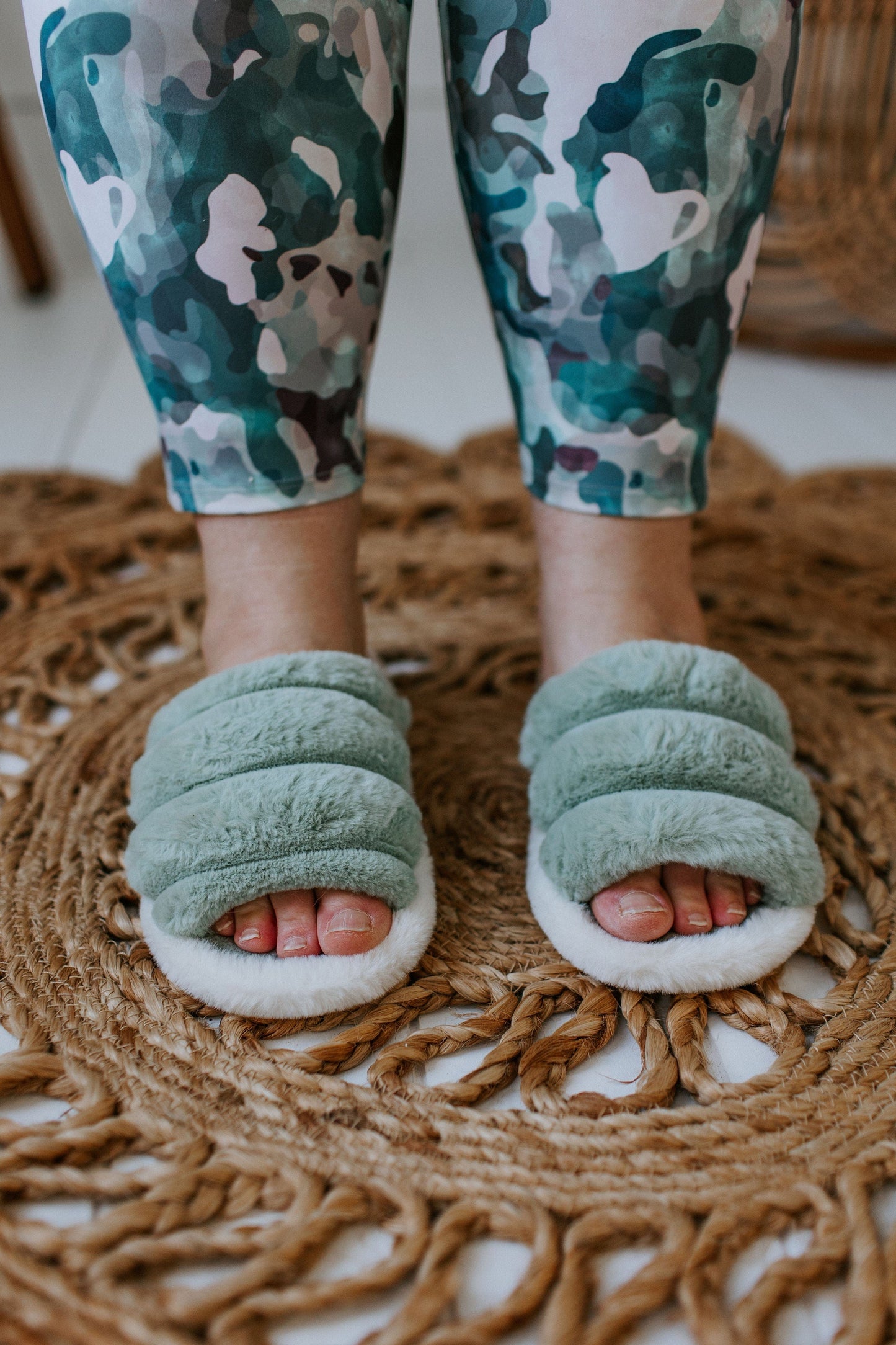 Close-up of feet in green plush slippers with rubber soles on a woven rug. The person is wearing camouflage-patterned pants.